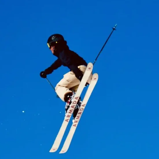 A skier in a black jacket and cream pants in the air against a blue sky.