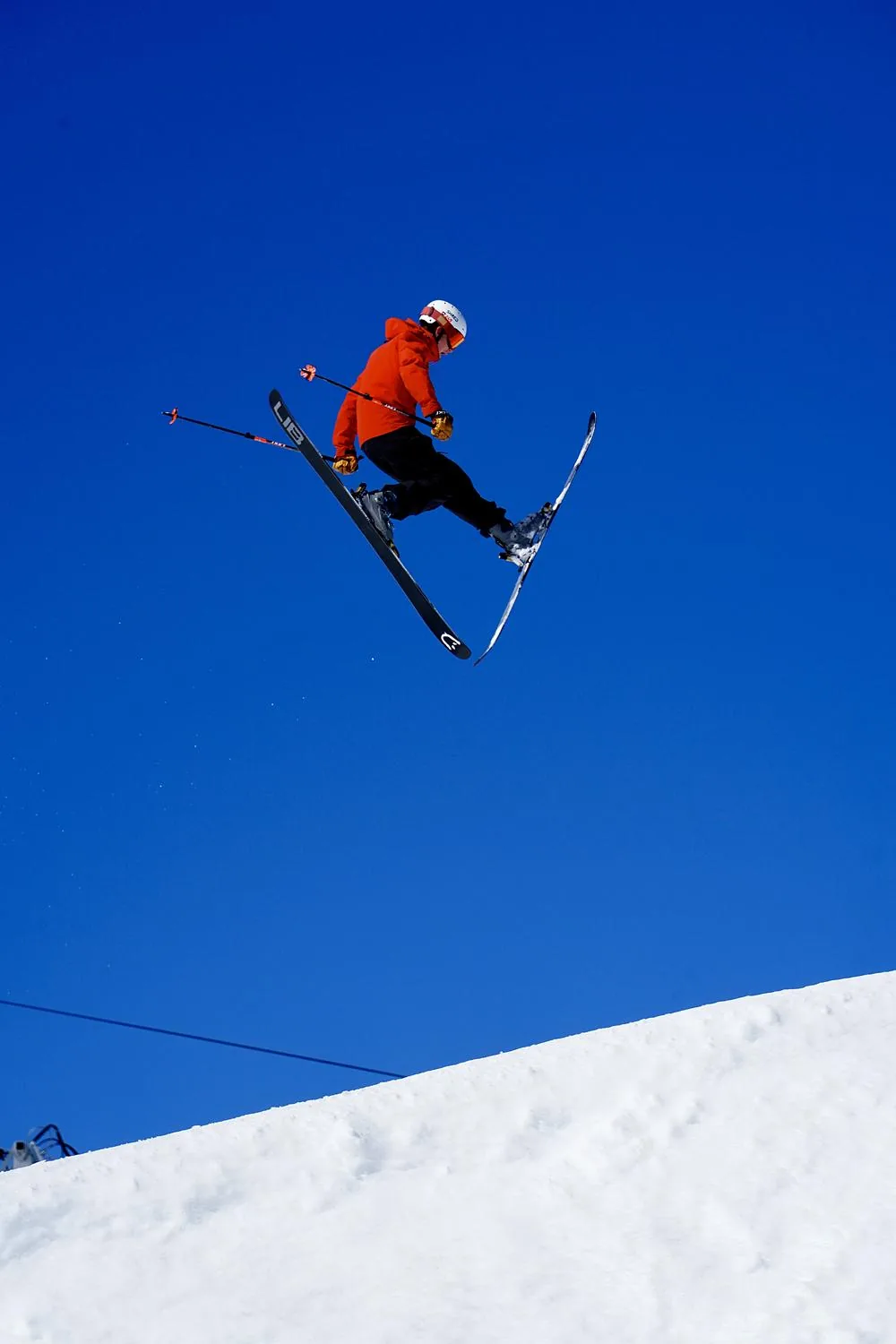 A skier in a red jacket airborne against a deep blue sky, the snow edge falling away below.
